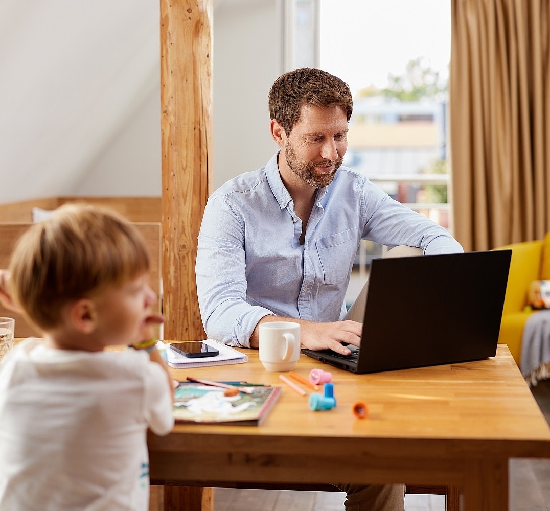 Ein Mann sitzt zuhause im Homeoffice an seinem Laptop. Bei ihm am Tisch spielen Kinder