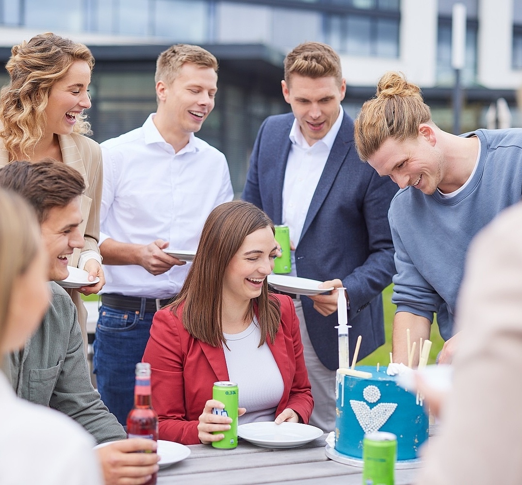 Lächelnde Mitarbeiter stehen und sitzen um einen Tisch und essen Kuchen 