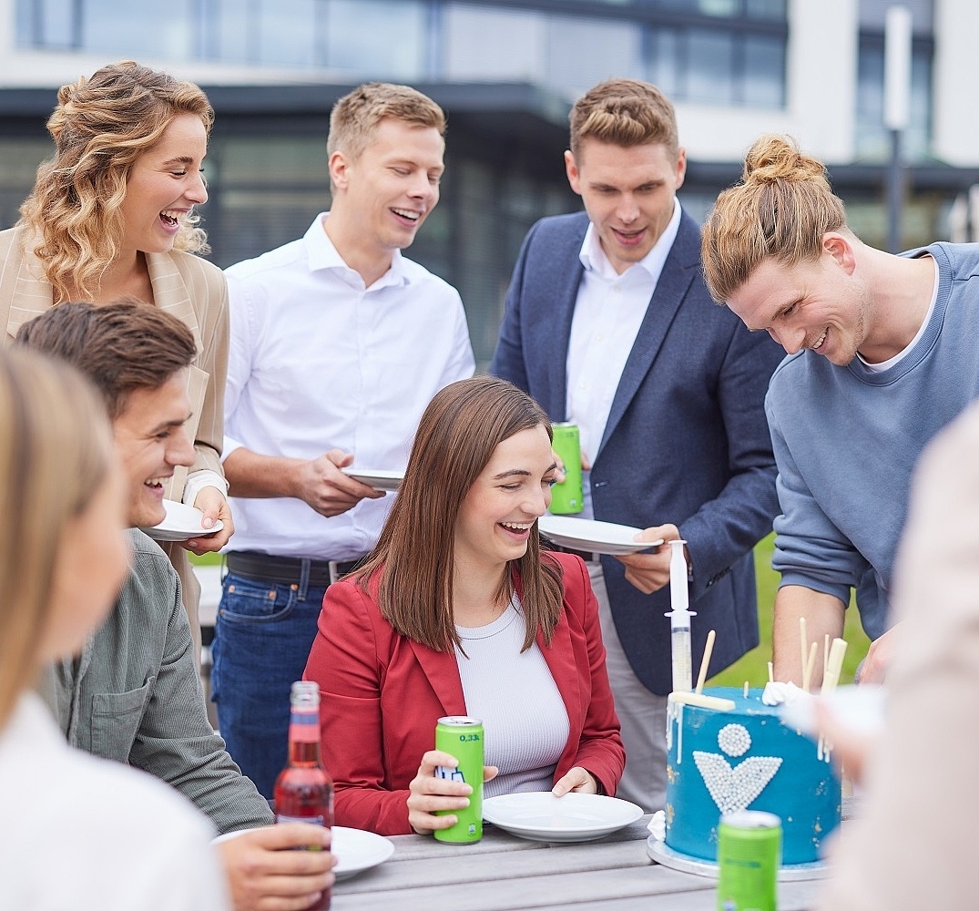 Lächelnde Mitarbeiter stehen und sitzen um einen Tisch und essen Kuchen