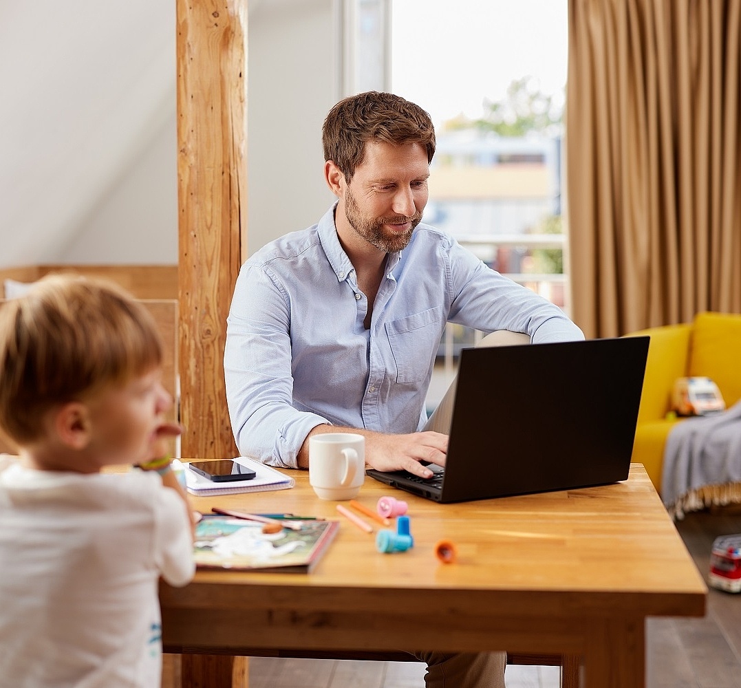 Ein Mann sitzt zuhause im Homeoffice an seinem Laptop. Bei ihm am Tisch spielen Kinder 