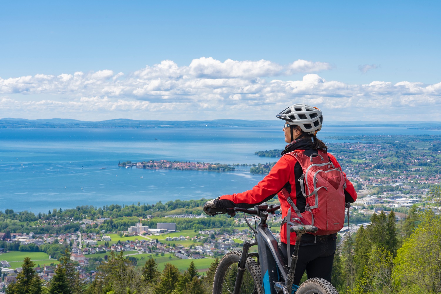 Eine Mountainbike-Fahrerin genießt das Alpenpanorama und blickt auf den Bodensee