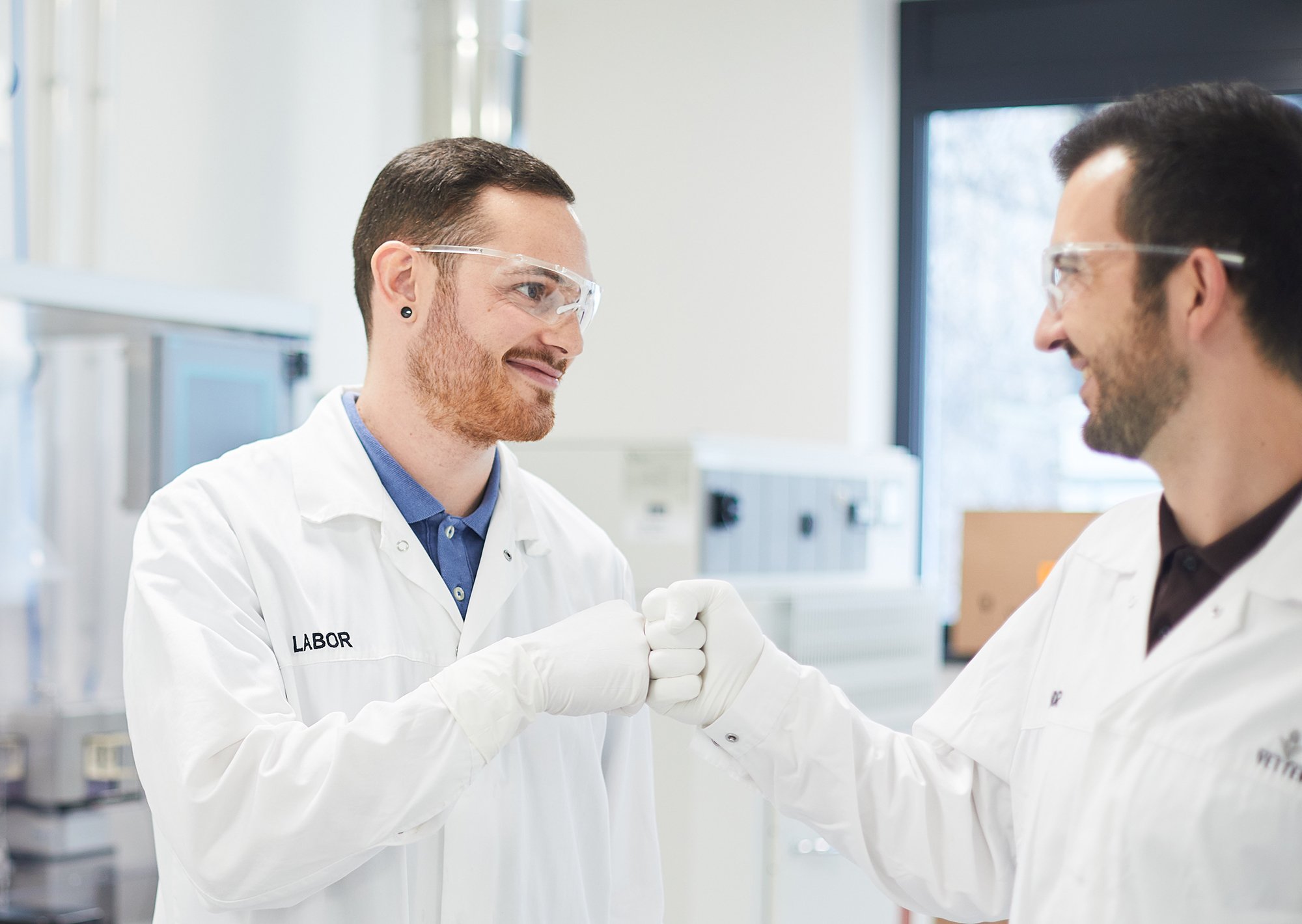 Zwei Mitarbeitende in Laborkleidung lächelnd im Labor. Die zwei geben sich einen Faustcheck 